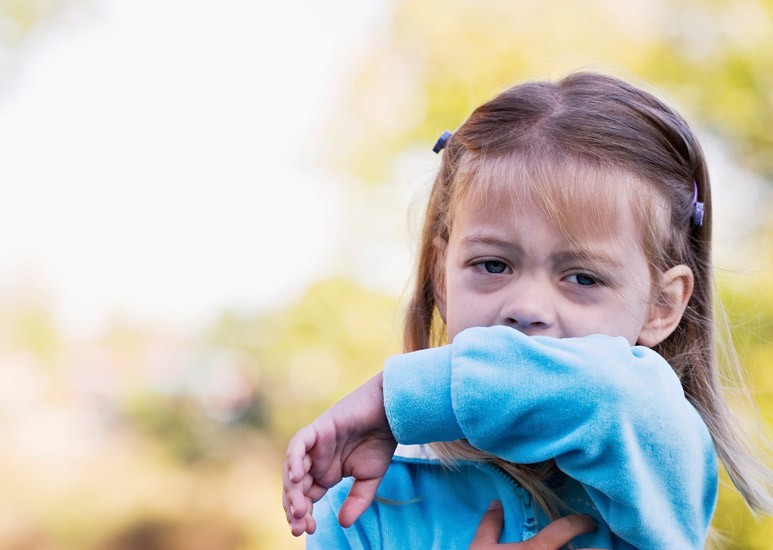 A young girl in a blue sweater coughs into her arm outdoors