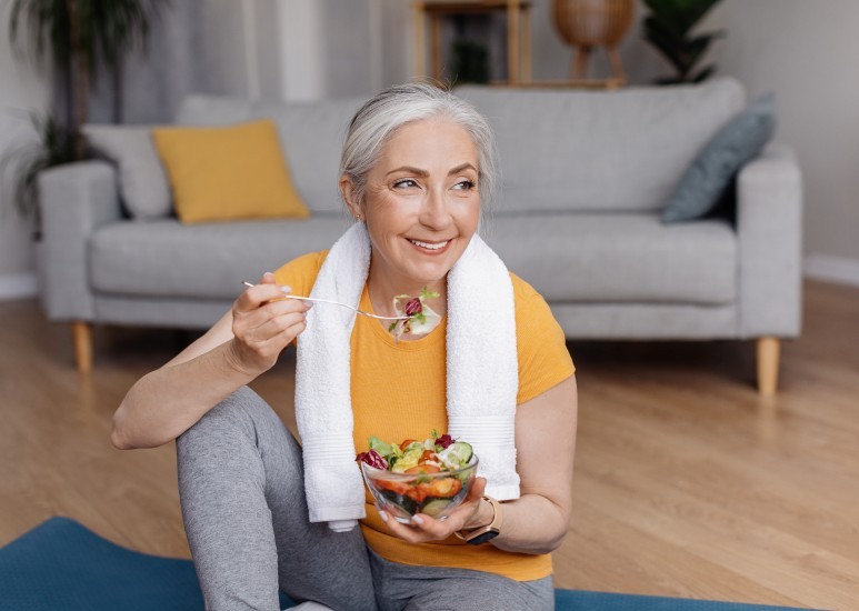 Woman eating a fresh salad after a workout while sitting on a yoga mat at home.