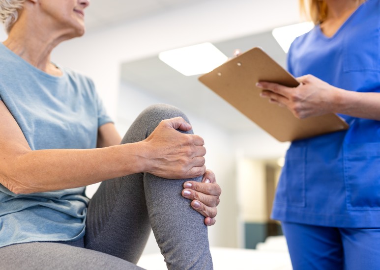  Senior woman holding her painful knee while a professional female therapist with a clipboard assesses her joint pain and mobility