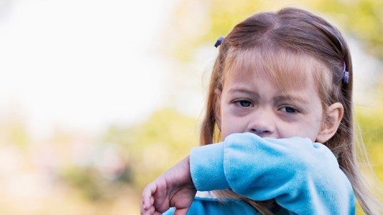 A young girl in a blue sweater coughs into her arm outdoors
