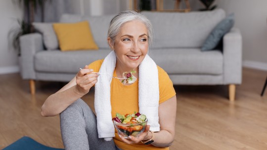 Woman eating a fresh salad after a workout while sitting on a yoga mat at home.