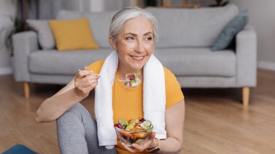 Woman eating a fresh salad after a workout while sitting on a yoga mat at home.