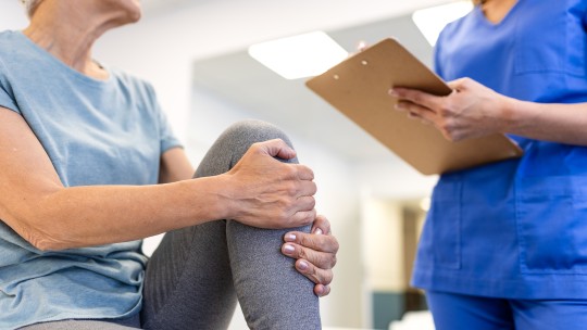  Senior woman holding her painful knee while a professional female therapist with a clipboard assesses her joint pain and mobility